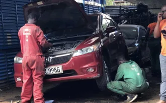 Two mechanics in a Nairobi garage inspecting a red Honda vehicle, one checking the engine and another working on the front wheel.