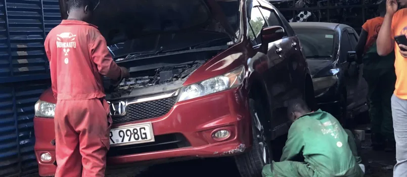 Two mechanics in a Nairobi garage inspecting a red Honda vehicle, one checking the engine and another working on the front wheel.