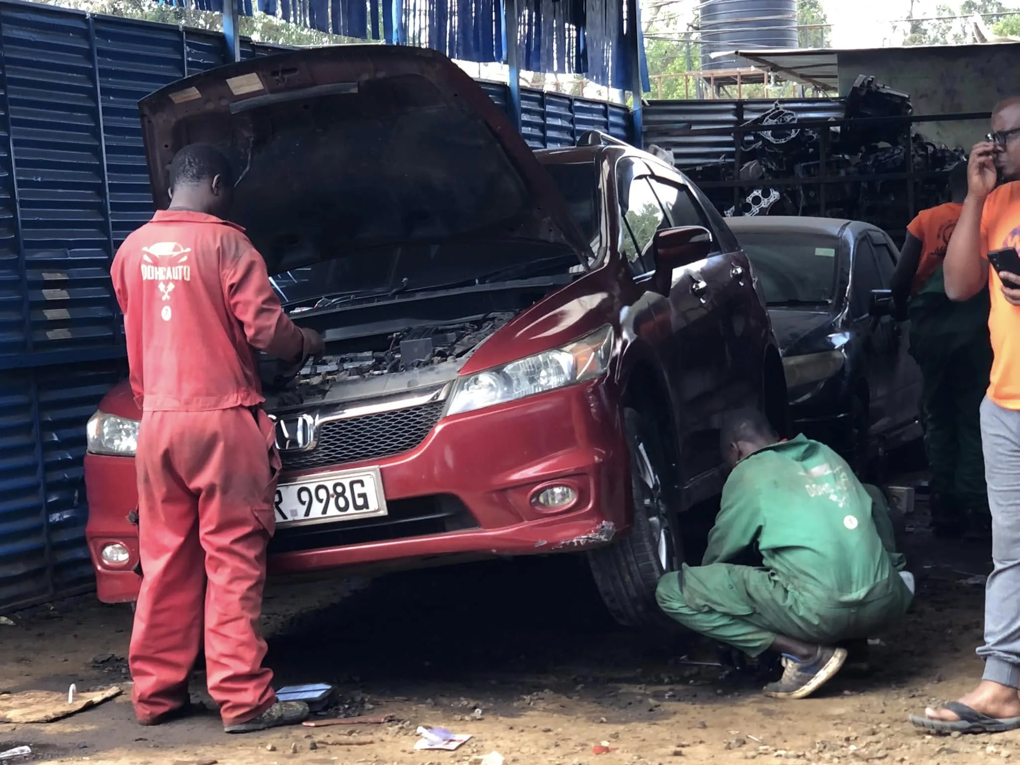 Two mechanics in a Nairobi garage inspecting a red Honda vehicle, one checking the engine and another working on the front wheel.